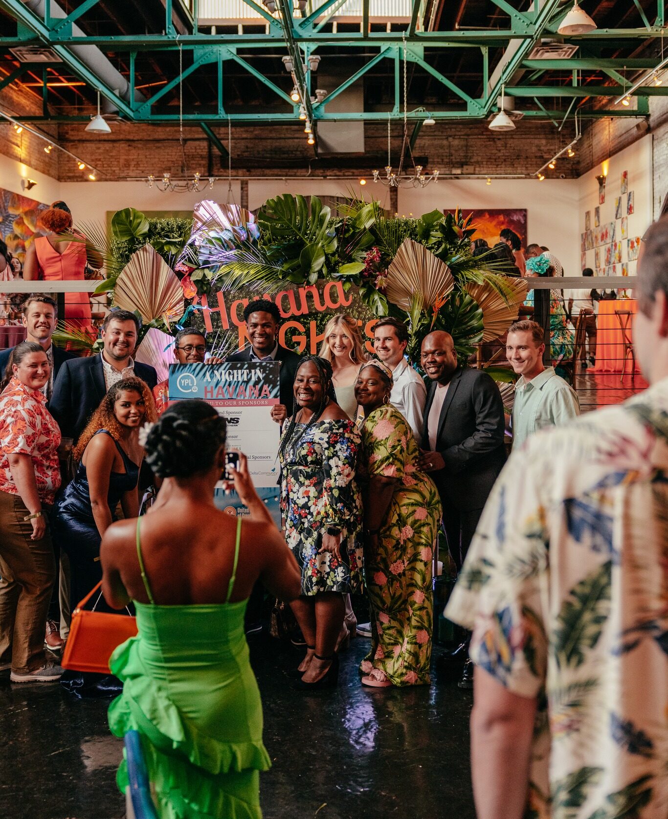 Attendees of "A Night in Havana" in festive and brightly patterned attire pose in front of a backdrop of tropical plants.